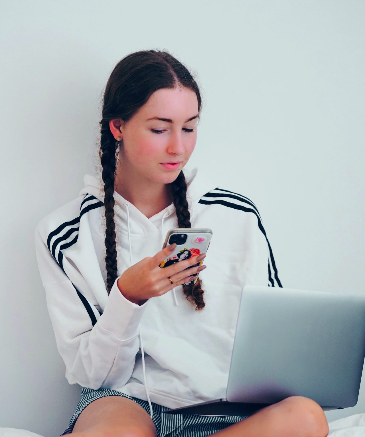 Eine Jugendliche sitzt auf dem Bett und schaut aufs Handy, während sie einen Laptop auf dem Schoß hat.
